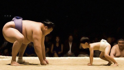 A man and a boy are on all fours, about to sumo wrestle each other.