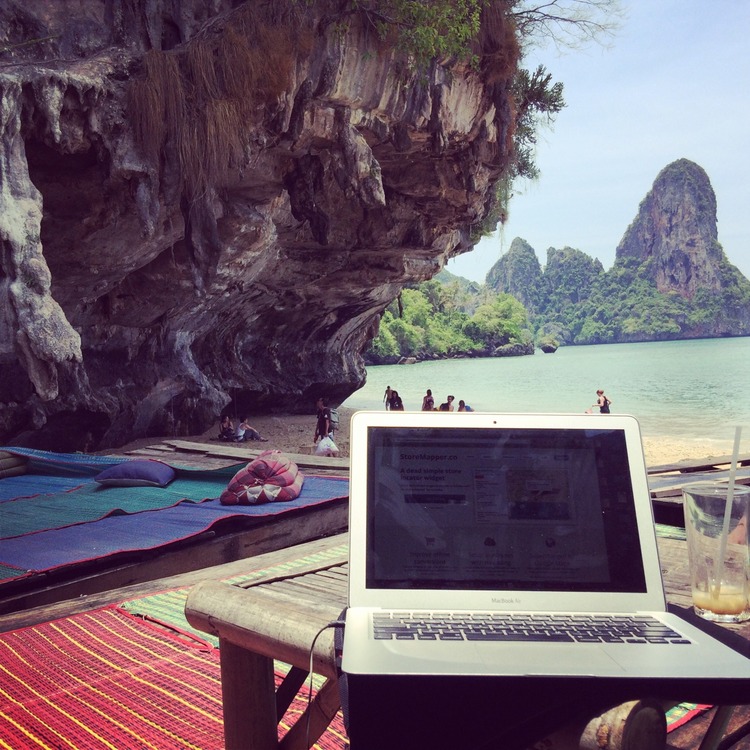A MacBook Air and an empty glass on a wooden table at a gorgeous beach with cliffs, trees, and clear water in the background.