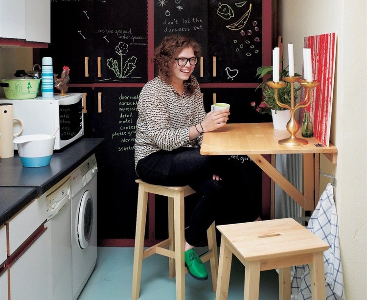 A woman is sitting on a wooden stool and holding a coffee mug in her hand in a home office that doubles as a kitchen with a wall-mounted table.