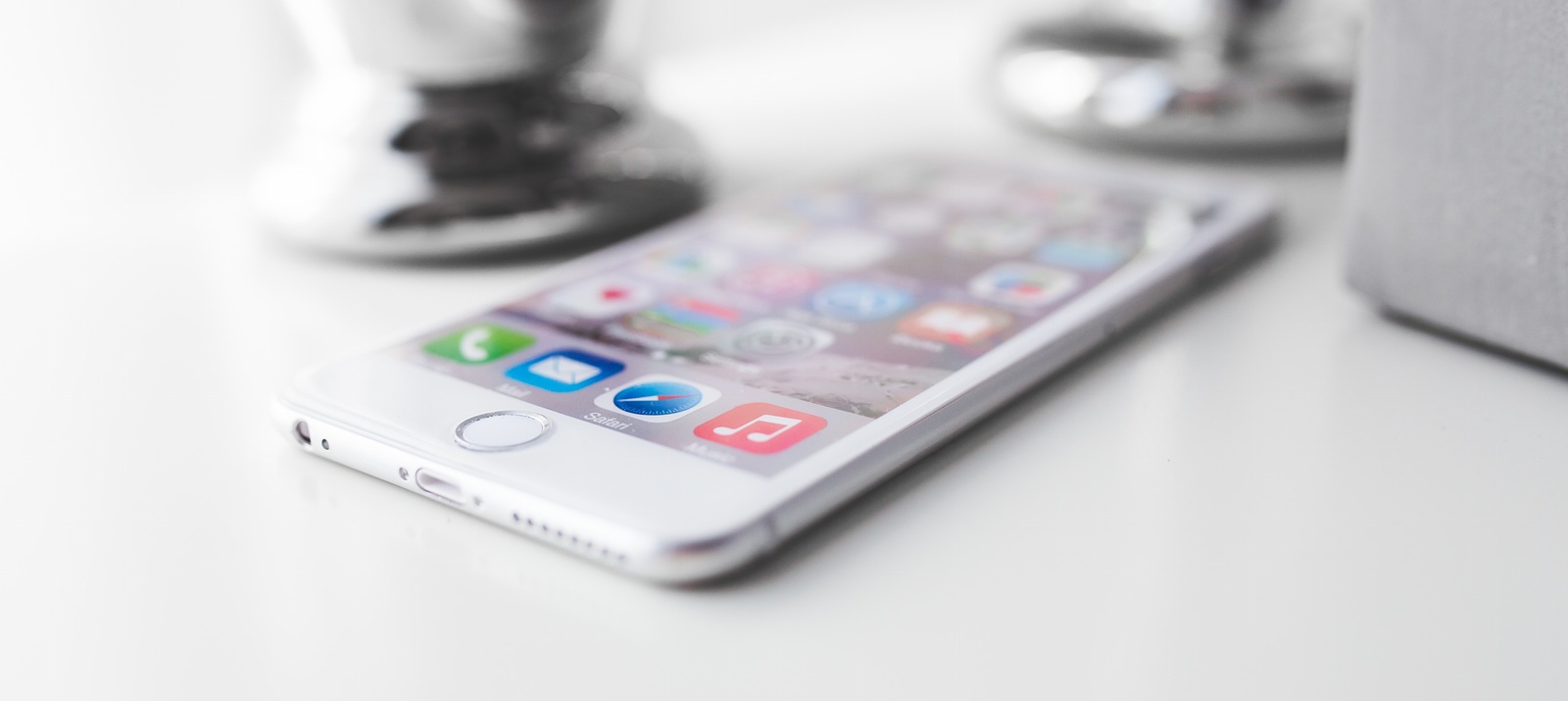 A white Apple iPhone 6 shows its homescreen apps while on a white table.
